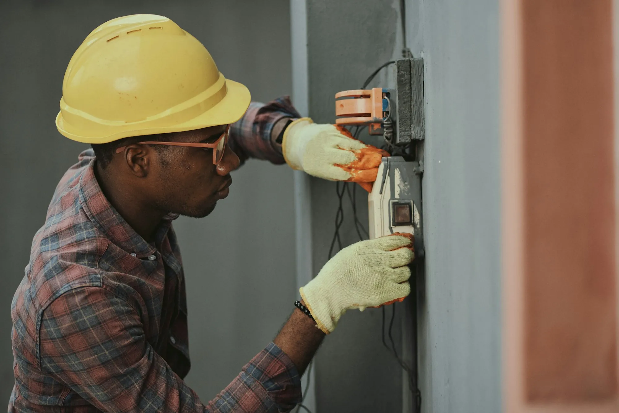 Electrician wearing a yellow helmet and gloves at a wall-mounted meter box